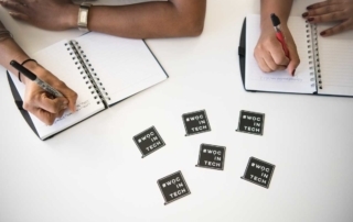 Aerial view of a table on which two pairs of hands are seen writing notes in notebooks, in front of them are six stickers that each say #WOC in Tech (women of color)